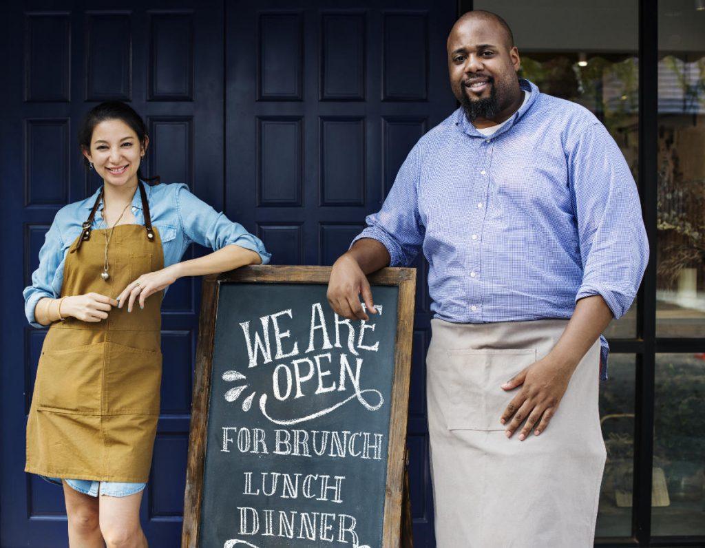 Two business owners, one male and one female, stand next to an open blackboard displaying information about their business opening hours and offering