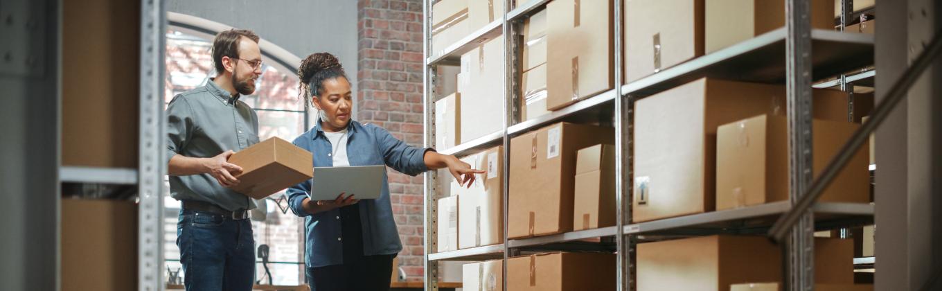 A male and female in the doorway of a storage room carrying a box to place on a shelving unit