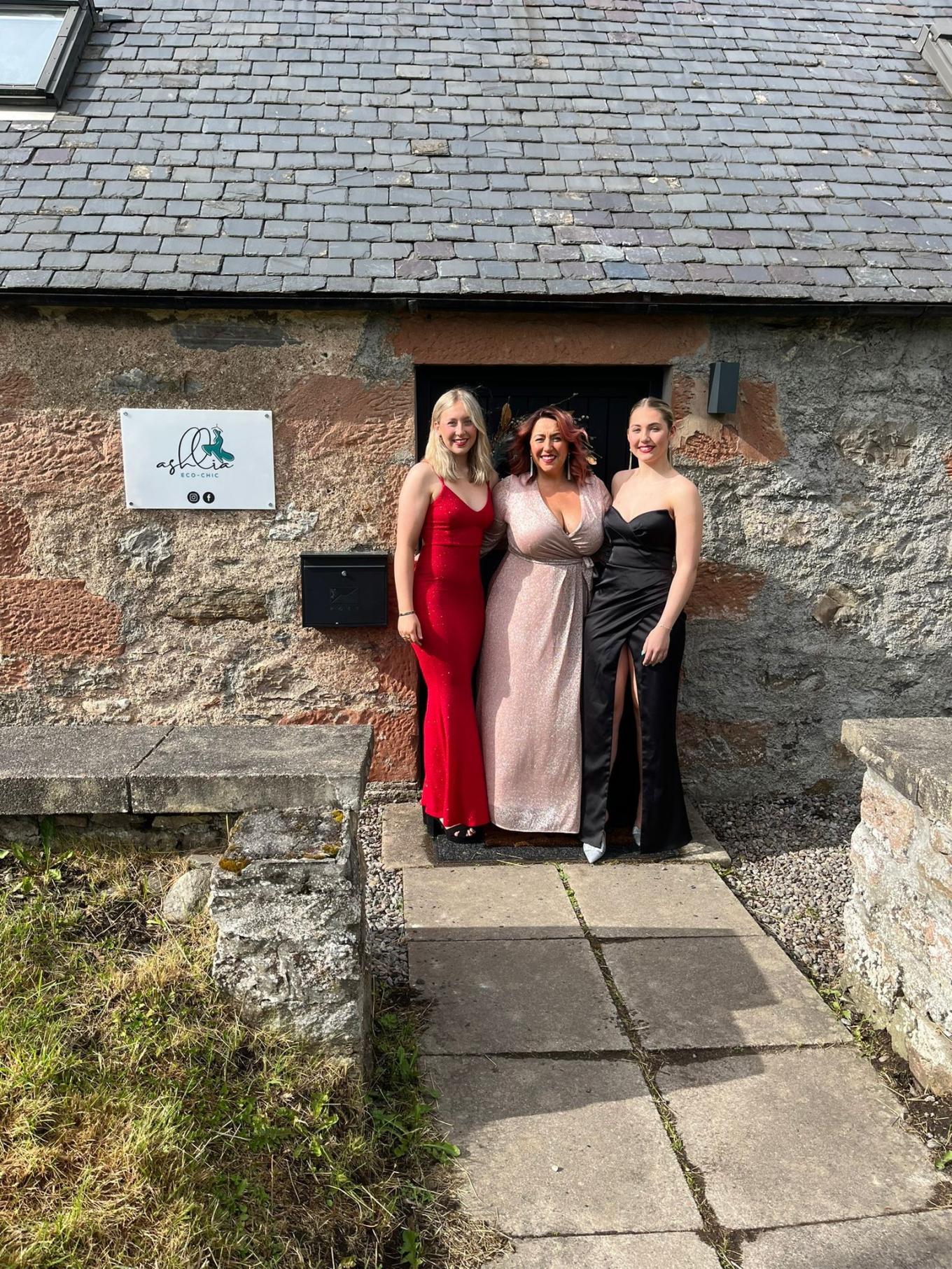 Three women are standing side by side outside a stone building with a sign on the wall. They are wearing long dresses in red, pink, and black.