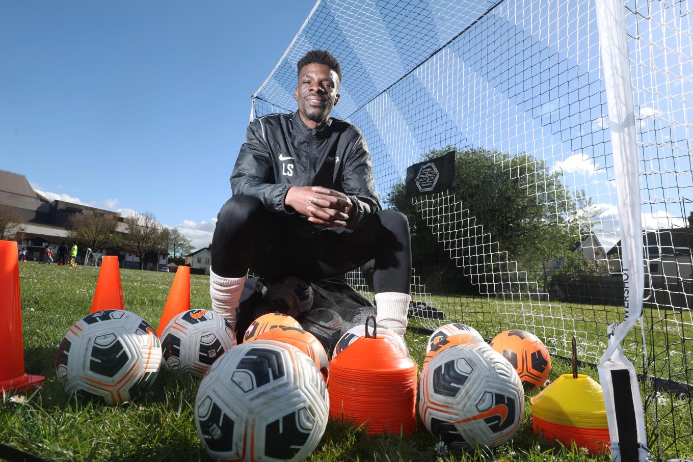 Founder of We Make Footballers, Leon Stapleton kneeling in front of a football goal surrounded by footballs and equipment