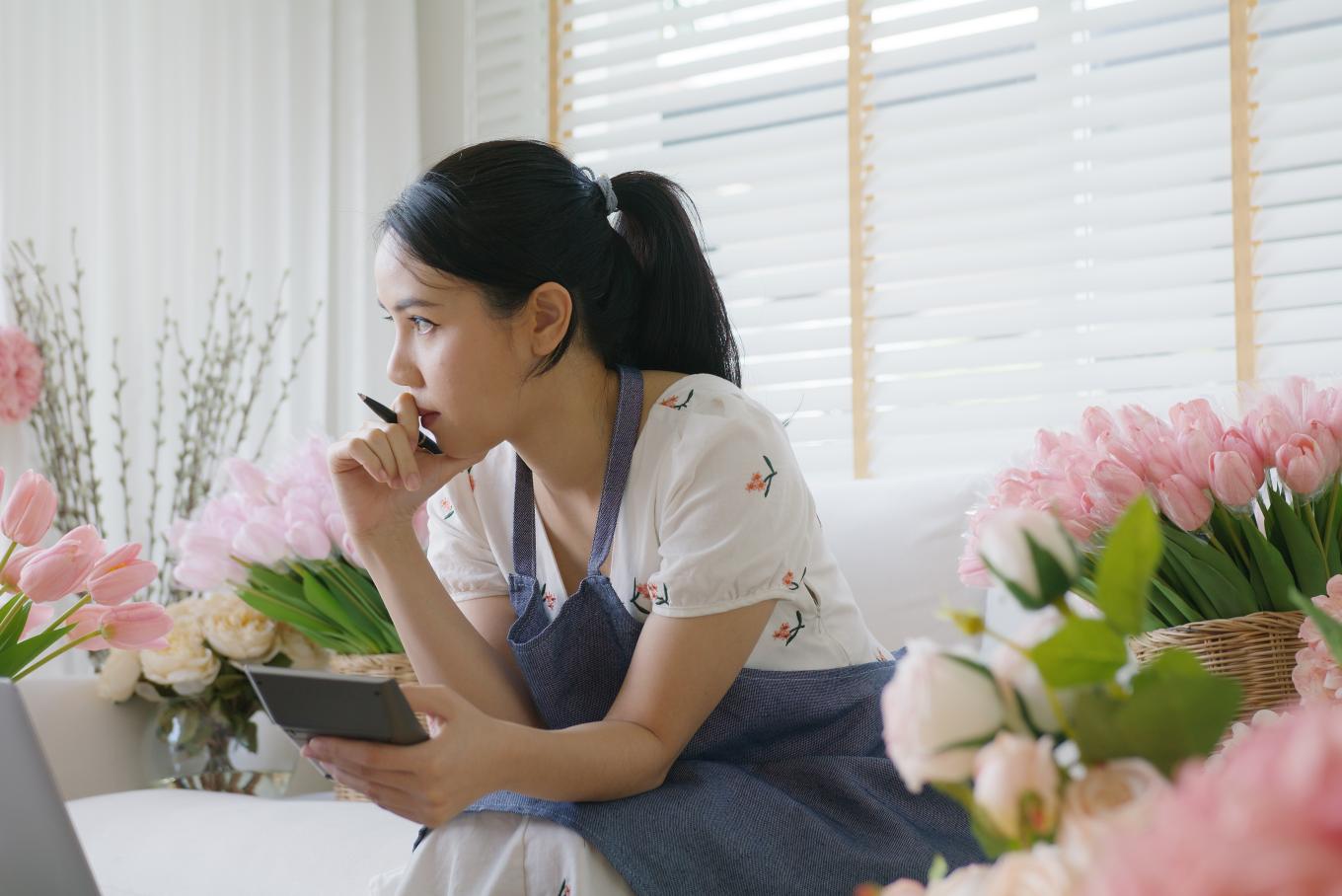 A female florist, sat in thought looking holding a calculator