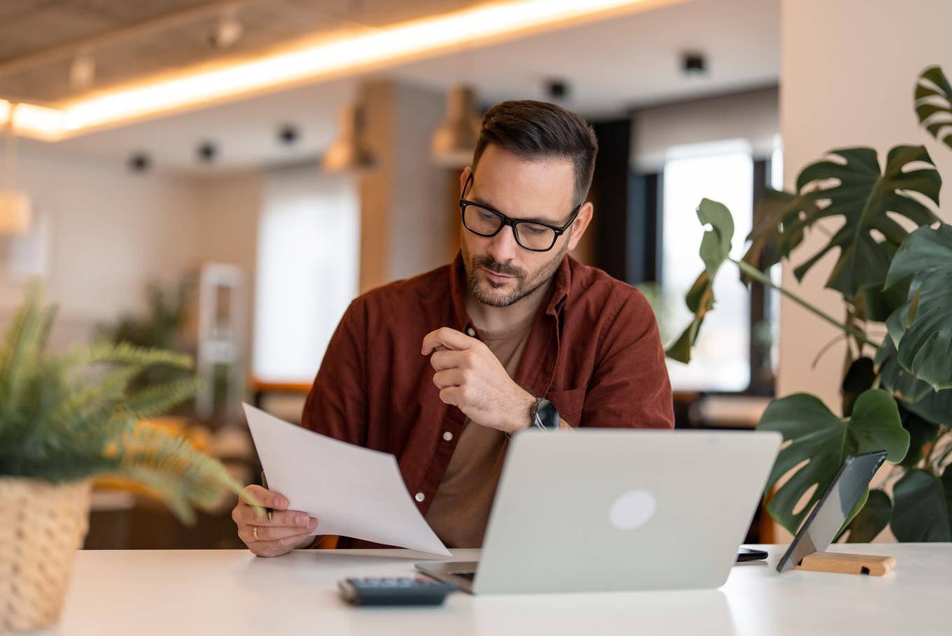 A young man wearing glasses, sat at a coffee shop bar at a laptop and holding a peice of paper 