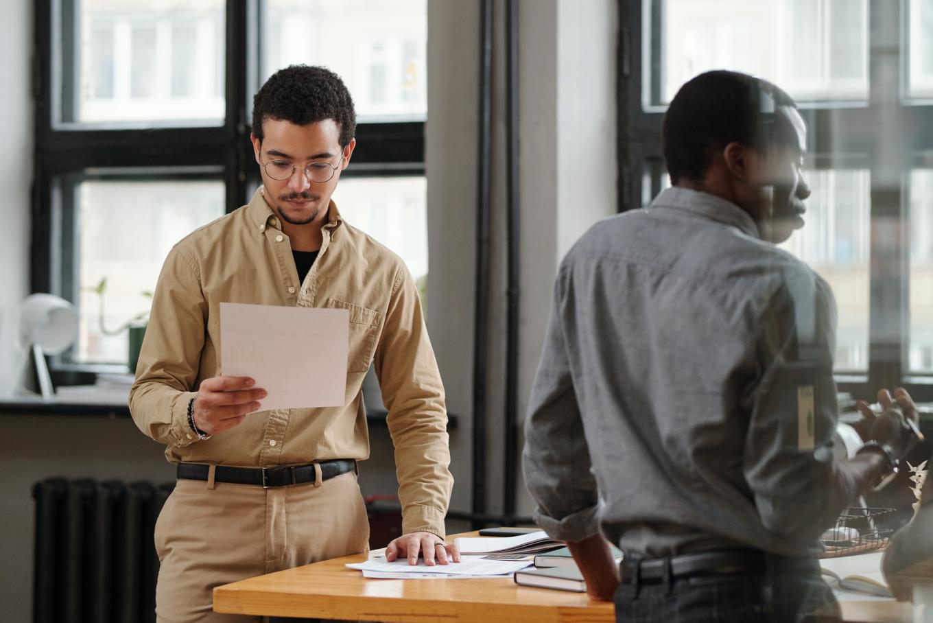 Two males standing in an office environment, one with their back turned the other reading a document