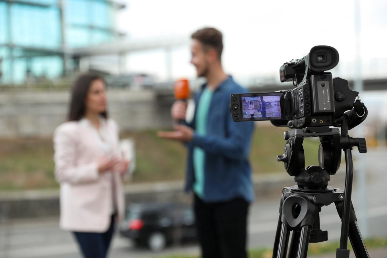 A blurred image of a male reporting interviewing a woman with a camera in the near distance in focus.